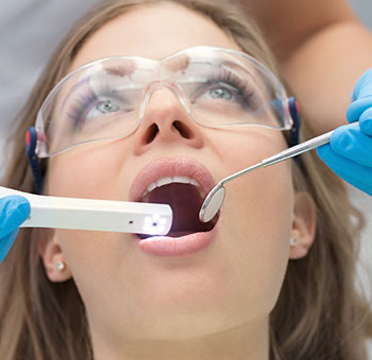 The image shows a woman receiving dental treatment, with a dental professional using a device to examine her mouth while she is seated in a dental chair. She appears to be undergoing a procedure that involves the use of a dental mirror and light, possibly for cleaning or examination purposes.
