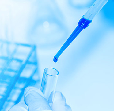 An image of a hand holding a small blue vial over a clear glass beaker filled with a blue liquid, set against a background featuring scientific equipment and a person's hand wearing gloves.
